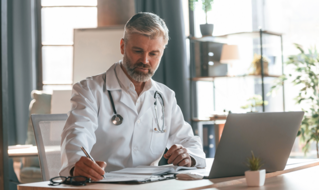 Doctor at a desk writing notes with a laptop in front of him